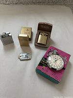 The full lot displayed on a white surface including three lighters, a flint packet, and a stopwatch with box.