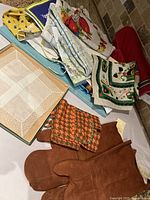 Top view of assortment of vintage kitchen linens including tea towels, lace handkerchiefs, placemats and a pair of leather oven mitts.