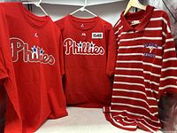 Front view of three Phillies shirts hanging; two t-shirts and one red and white striped polo shirt