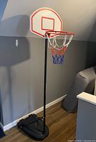 Full view of the portable basketball hoop showing the black adjustable pole, base, white backboard with red markings, and orange rim with a three-colored net.