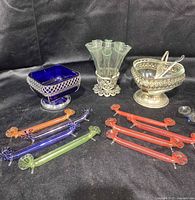 Antique ornate serving pieces displayed, featuring cobalt blue relish dish, clear glass bud vase, ornate metal bowl, striped glass flatware holders in red, blue, purple, and green, and clear glass stirrers.