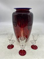 Photo of red art glass vase with blue rim and three clear cordial glasses with red bases all displayed together.