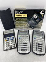 Sharp OZ-7620 electronic organizer with original box and carrying case next to two TI-36X Solar calculators and a Casio Memory B-1 calculator shown front view.