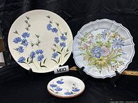 Three decorative floral plates shown on black display stands. One large 16-inch round plate with blue flowers, one medium 14-inch scalloped plate with multicolor flowers, one small 6-inch plate with blue flowers.