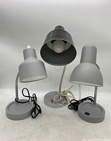 Three modern gooseneck desk lamps in a row on a white surface: two gray and one black lamp with metal flexible neck and circular bases.