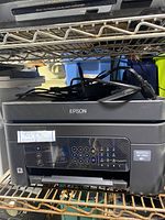 Front view of Epson Workforce WF-2950 printer showing keypad, display screen, and paper feed slot. Printer is black with the Epson logo visible on top.