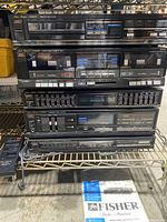 Front view of stacked Fisher stereo components including cassette decks, equalizer, amplifier, and CD player, with remote control on side and instruction manual at base.