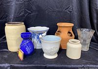 Six assorted flower pots and a clear glass vase arranged side by side on black background.