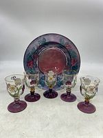 Front view of five hand-painted cordial glasses in a row with one glass plate behind them, showing purple stems and colorful floral patterns on glass.