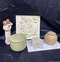 Five home decor items displayed against a black fabric background including a figurine sculpture, two lidded containers, a carved stone sphere on marble base, and a floral relief plaque.