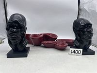 Front and side views of two black African bust statues displayed on bases alongside three maroon ceramic bowls with an interconnected design.