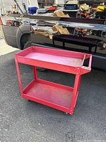 Full view of the red metal shop cart showing two shelves and handlebar, placed on asphalt beside a vehicle.
