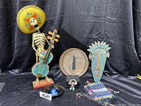 Display of Day of the Dead skeleton figurine, pottery plate, copper tribal mask, small woven rugs, and other small items on black background.