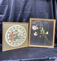 Two framed needlepoint artworks displayed against a dark backdrop showing the rose floral and Asian scene designs.