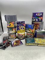 Wide shot showing multiple unopened Memorex 25-pack CD-R spindles, packs of CD/DVD sleeves, and a stacked group of empty jewel cases.