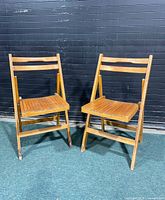 Pair of wooden folding chairs with slatted seats and backs, positioned on a carpeted floor against a black paneled background.