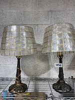 Pair of two table lamps approximately 24 inches tall with mosaic glass square panel shades and metal ornate bases on a metal shelf against a concrete block wall.