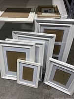 Photo showing nine empty white photo frames arranged on the floor, varying sizes and styles with some wide borders.