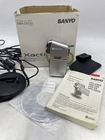 Sanyo Xacti C5 camera placed next to instruction manual, black carrying case, docking station, and original white box with Sanyo branding.