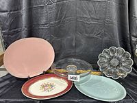 Four serving platters arranged against a black fabric background: pink oval plate, clear glass divided platter, white plate with maroon floral rim and flower design, light blue oval platter