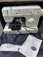 White Singer electronic control sewing machine in open wheeled carrying case with motor and foot pedal on base. Includes manual and paperwork.