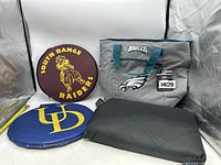 Photo of all four items including the Eagles cooler bag, the two round seat cushions with school logos, and the black waterproof foldable blanket.