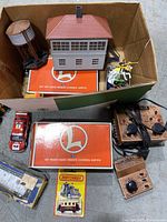 Box full of various Lionel toy train parts including two orange and white remote-control switches, plastic station building, water tower, several small toy train cars and figurines.