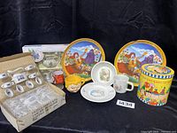 Full view of all vintage children's dish sets and collectibles arranged on black background showing variety of items including plates, spoons, cups, glass and tin.