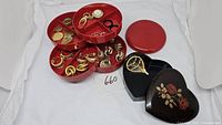 Red tiered round jewelry box open showing assortment of gold and silver-tone earrings, rings, pendants, and keychain with watch and pearl detail; next to a closed red lid and black heart-shaped jewelry box with floral rose design containing gold-tone brooch shaped like hot air balloon.