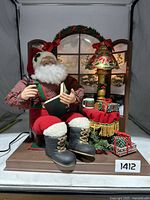 Front view of vintage Santa Claus reading figurine with Christmas decorations including gifts, lamp, wreath, and mug on wooden base.