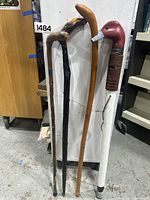 Four walking sticks leaning against a board, showing different handle styles including duck head, curved plain wood, snake carving, and plain straight black wood.