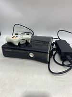 Xbox 360 console with white controller on top and power supply beside it, showing front and top side of the console.