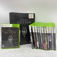 Front view of The Elder Scrolls V Skyrim game case, Skyrim official guide book, and 11 Xbox 360 game cases upright in a row.