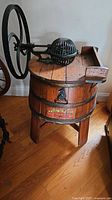 Full view of the antique wooden barrel washing machine with black metal wringer and large hand crank wheel on a hardwood floor.