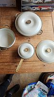 Three vintage Copco cookware pieces on wooden floor: two round pots with lids and dual handles, one saucepan with lid and single long handle, cream colored with brass handles and knobs.