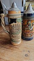 Two German beer steins on wooden surface, showing detailed artwork and pewter lids.
