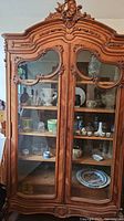 Frontal view of large ornate wooden china display cabinet showing carved details and glass paneled doors, with contents inside.