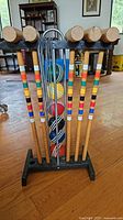 Front view of vintage croquet set showing 5 wooden mallets with color bands hanging on black wooden stand and 6 colored balls stacked vertically with metal wickets behind balls.