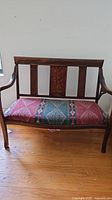 Wooden 2-seat bench with floral design on backrest and multicolor fabric seat upholstery, shown on wooden floor.