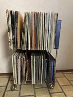 Front view of two levels of vinyl records tightly packed on a metal rolling stand showing many records with visible covers and spines.