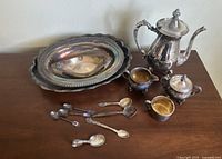 Full lot group showing tea set, serving trays, and collection of spoons on brown surface against wall.