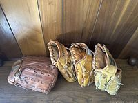 Four vintage baseball mitt gloves arranged side by side on a wood surface with a wood panel background.