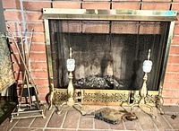 Front view of brass and ceramic andirons, brass fire dogs, and metal log holder arranged in front of brick fireplace with brass screen.