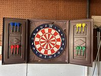Full frontal view of vintage Champion dart board with open brown case and 12 darts in four colors mounted in holders on case doors and board center showing significant vintage wear.