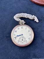 Front dial view of the vintage Elgin pocket watch showing white dial, black numerals, blue hands, and braided fabric loop attached.