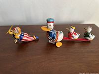 Photo showing all three tin wind-up toys together: a patriotic clown and a duck family with two smaller ducklings attached by a red bar.