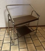 Side angle photo of vintage bar trolley showing metal frame, two wooden shelves and caster wheels.