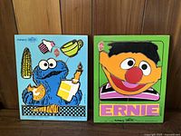 Two vintage Sesame Street wooden puzzles side by side on a wooden floor against a wood-paneled wall. Left is the Cookie Monster puzzle with multiple pieces and colorful images of corn, cookie, candle, cup, and carrot. Right is the Ernie puzzle with the character's face and clothing in bright colors.