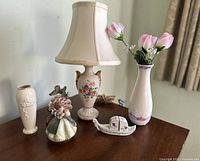 Photo of the lot showing vintage porcelain lamp, tall vase with pink tulip flowers, smaller floral vase, girl figurine, butterfly figurine, and boat-shaped dish on a wooden table.
