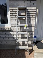 Front view of silver aluminum step ladder with 6 steps leaning against white brick wall under bright daylight.
